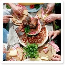 Eine Gruppe von Menschen, die um einen Holztisch herumsitzen, mit verschiedenen Lebensmitteln, einschließlich Fleisch, Käse und Brot, auf Holzbrettern angeordnet. Der Tisch ist mit einem weiß-rot karierten Tuch bedeckt.