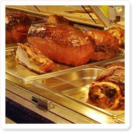 Ein Buffettablett mit verschiedenen Fleischsorten, darunter gebratene Ente, Schwein und ein ganzer Braten, der mit Glanz überzogen ist. Das Tablett ist aus Edelstahl mit einer silbernen Oberfläche und auf einer glänzenden Unterlage platziert.