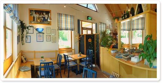 Ein gemütliches Interieur eines Restaurants mit Holzmöbeln, blauen Stühlen und einer gelben Theke. Einige Pflanzen und Bilderrahmen schmücken die Wände. Der Raum hat eine warme und einladende Atmosphäre.