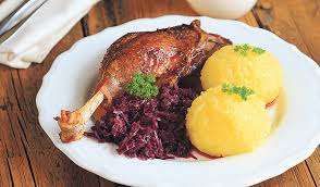 Ein weißer Teller mit gebratener Ente, Rotem Kohl und zwei gelben Knödeln, garniert mit Petersilie, steht auf einem Holztisch.