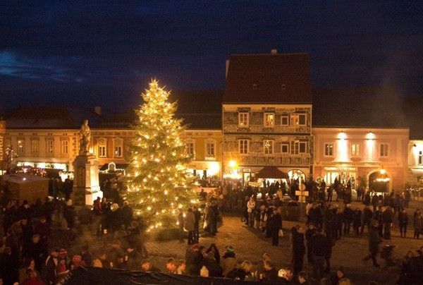Ein hell beleuchteter Weihnachtsbaum auf einem Stadtplatz mit vielen Menschen, die bei Nacht herumlaufen.