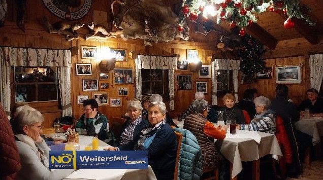 Eine Gruppe älterer Menschen versammelt sich in einem warm beleuchteten rustikalen Restaurant mit Holzwänden und gerahmten Fotos. Sie sitzen an Tischen mit Getränken und Essen, und ein Weihnachtsbaum und Dekorationen schmücken den Raum.