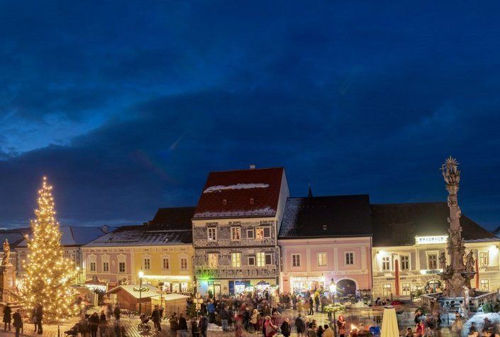 Eine lebhafte Abendmarktszene auf einem Stadtplatz mit Gebäuden, einem Weihnachtsbaum und vielen Menschen, die herumlaufen.