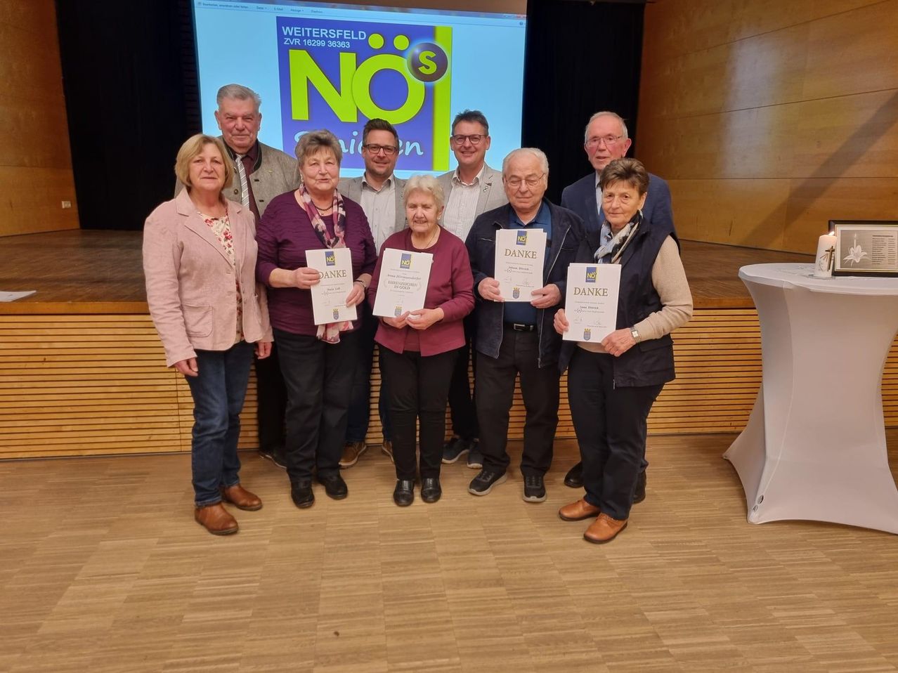 Eine Gruppe älterer Menschen mit Urkunden steht auf der Bühne. Eine Projektionsleinwand zeigt 'Weitersfeld ZVR 16299 38363 NO1'. Sie sind warm gekleidet und lächeln für ein Foto.