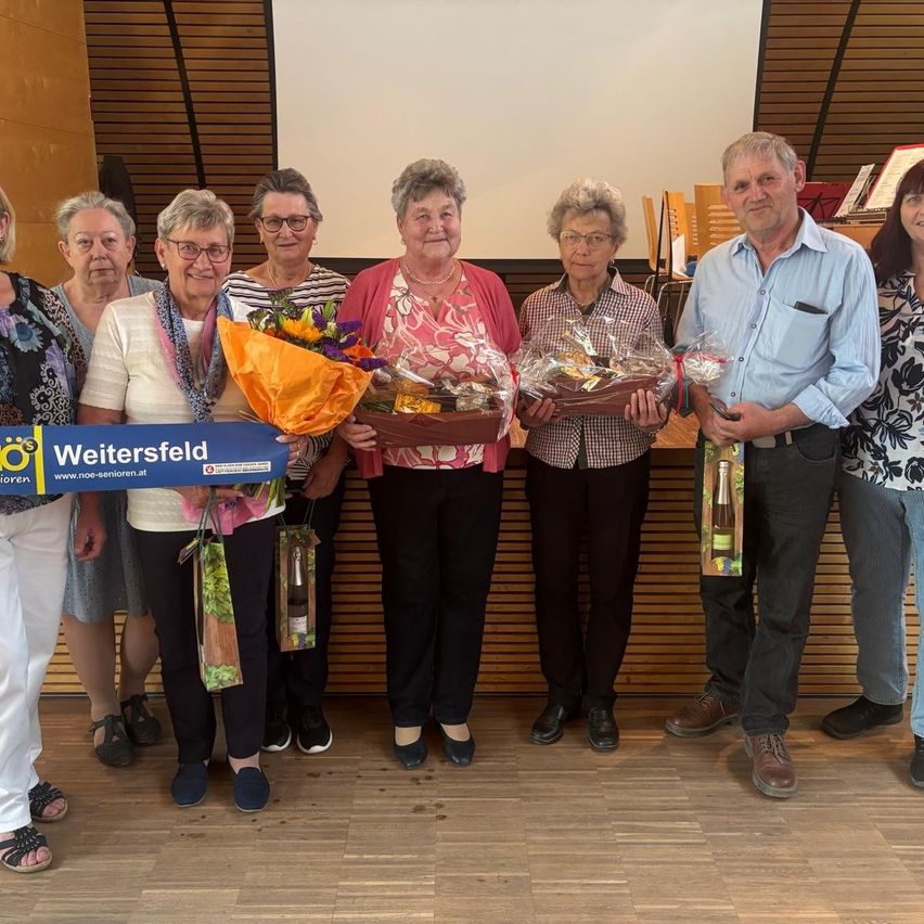 Eine Gruppe älterer Menschen steht zusammen und hält Blumen und Geschenke. Sie scheinen ein Ereignis zu feiern. Eine Frau hält ein blaues Schild mit dem Wort Weitersfeld. Hinter ihnen steht eine Holzwand mit einer weißen Leinwand.