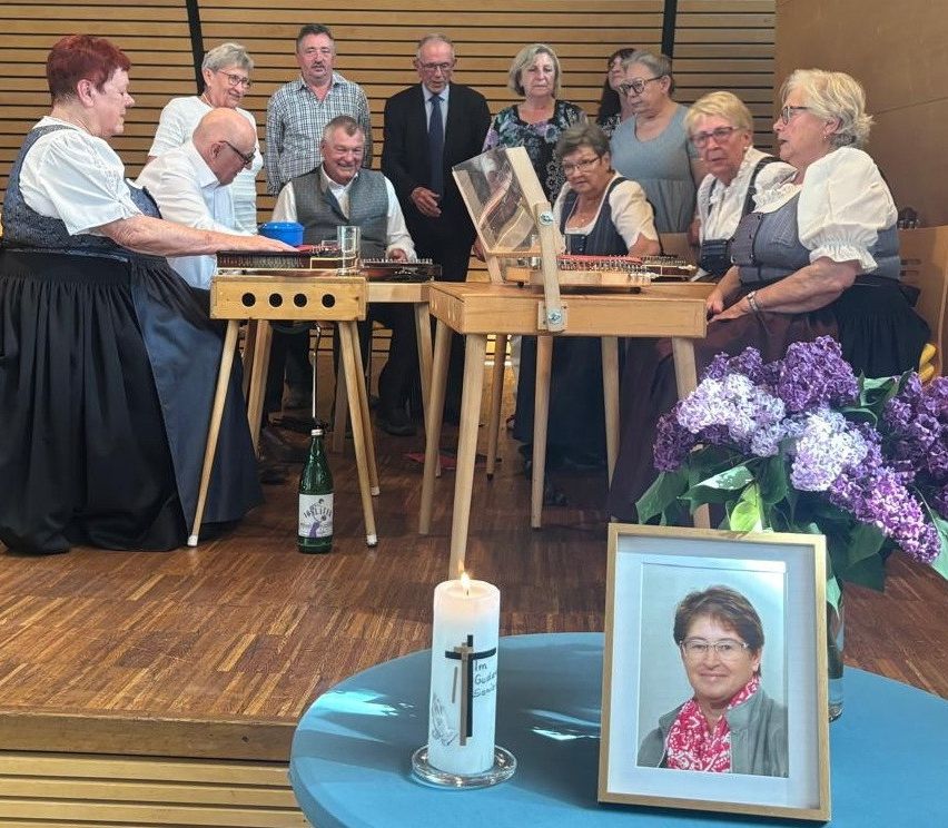 Eine Gruppe von Menschen spielt Musikinstrumente in einem Raum. Eine Kerze und ein gerahmtes Bild stehen auf dem Tisch.