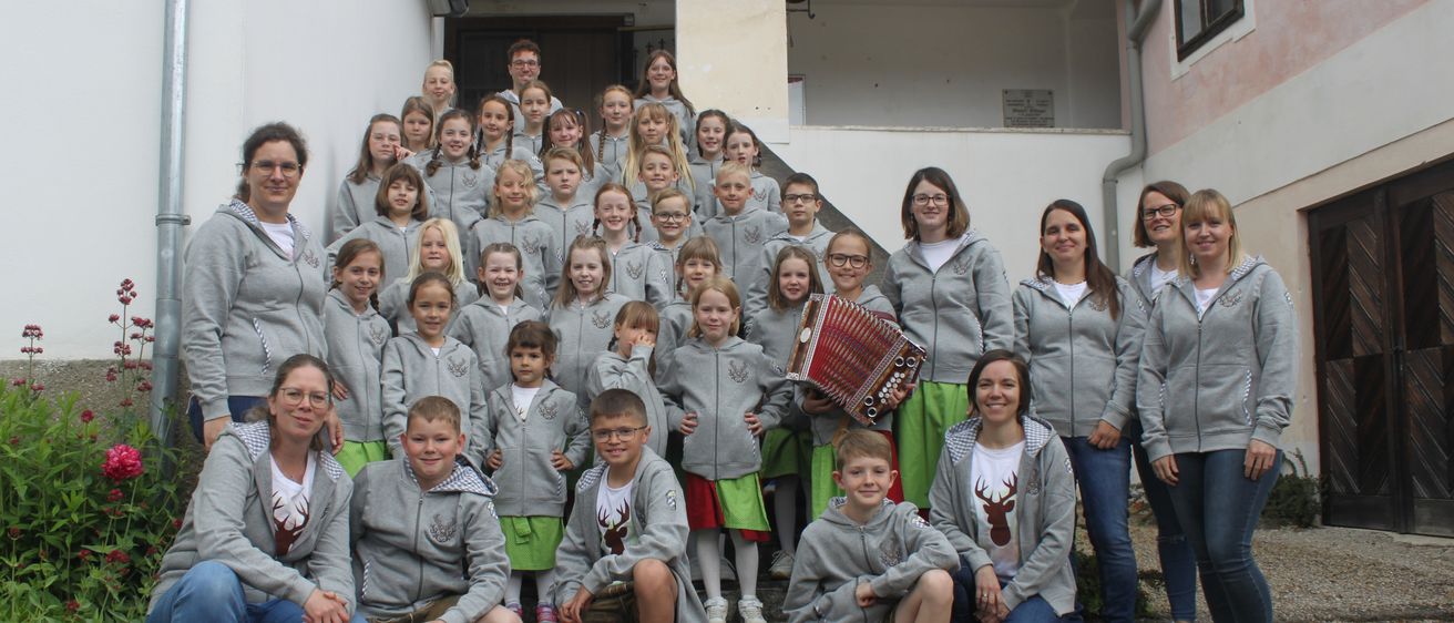 Eine Gruppe von Kindern und Erwachsenen, die graue Hoodies tragen, posiert für ein Foto vor einem Gebäude. Einige halten ein Akkordeon.