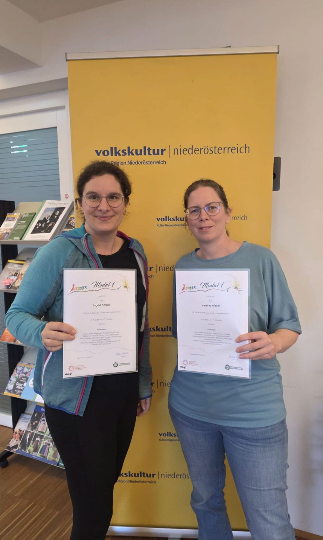Zwei Frauen stehen vor einem gelben Banner und halten Urkunden in der Hand. Sie tragen Freizeitkleidung. Die Frau auf der linken Seite hat eine Brille und eine blaue Jacke, während die Frau auf der rechten Seite ein graues T-Shirt trägt. Hinter ihnen befindet sich ein Bücherregal.