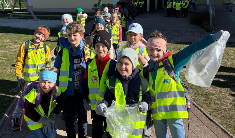 Eine Gruppe von Schulkindern in Sicherheitswesten steht in einer Reihe und hält Plastiktüten. Sie sind draußen auf einem gepflasterten Bereich.