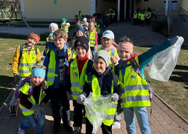Eine Gruppe von Schulkindern in Sicherheitswesten steht in einer Reihe und hält Plastiktüten. Sie sind draußen auf einem gepflasterten Bereich.