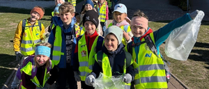 Eine Gruppe von Schulkindern in Sicherheitswesten steht in einer Reihe und hält Plastiktüten. Sie sind draußen auf einem gepflasterten Bereich.