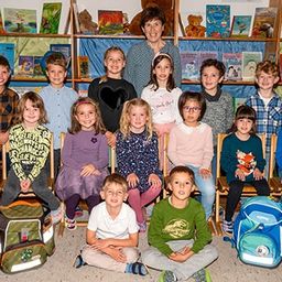 Eine Gruppe junger Kinder und ihre Lehrerin lächeln für ein Foto in einem Klassenzimmer mit Bücherregalen und Dekorationen.