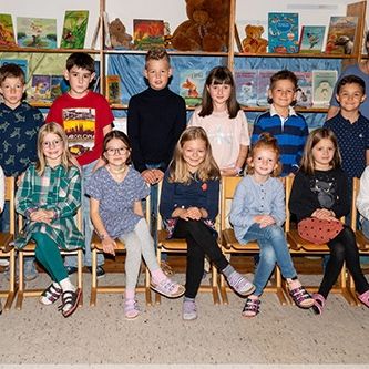 Eine Gruppe von Kindern und zwei Frauen sitzen in einem Klassenzimmer. Sie alle lächeln.