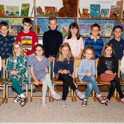 Eine Gruppe von Kindern und zwei Frauen sitzen in einem Klassenzimmer. Sie alle lächeln.