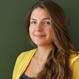 Eine Frau mit langen braunen Haaren und einem gelben Schal posiert lächelnd für ein Foto vor einer grünen Wand.