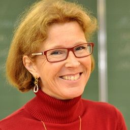 Eine ältere Frau mit kurzen blonden Haaren lächelt hell. Sie trägt rote Brille und ein rotes Rollkragenshirt. Sie steht vor einer grünen Tafel.