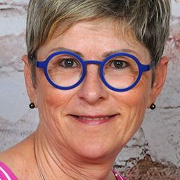 Eine Frau mit kurzen Haaren und blauen Gläsern lächelt vor einer Backsteinwand.