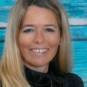 Eine Frau mit blonden Haaren lächelt und steht vor einem blauen Hintergrund. Sie trägt ein schwarzes Rollkragenshirt und eine Halskette.