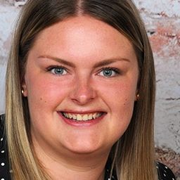 Eine Frau mit blondem Haar, die ein Shirt mit Punkten trägt, lächelt mit sichtbaren Zähnen vor einer Ziegelsteinwand.