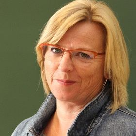 Eine Frau mit blondem Haar, orangefarbener Brille und Jeansjacke steht vor einem grünen Hintergrund und lächelt in die Kamera.