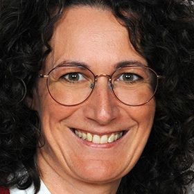 Eine lächelnde Frau mit lockigem schwarzem Haar und Brille. Sie trägt ein rotes Oberteil und scheint für ein Foto zu posieren.