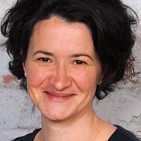 Eine lächelnde Frau mit kurzen schwarzen Haaren steht vor einer Ziegelmauer, sie trägt ein schwarzes Shirt. Sie hat einen warmen Ausdruck und scheint in die Kamera zu schauen.