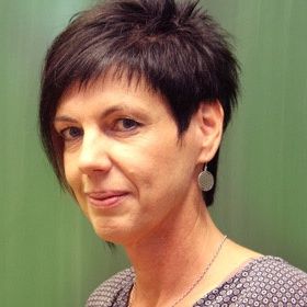 Eine Frau mit kurzen, schwarzen Haaren, die in die Kamera lächelt, trägt eine Halskette und Ohrringe. Sie steht vor einer grünen Wand.