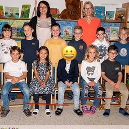 Eine Gruppe von Kindern, die lächeln, sitzen auf Stühlen in einem Klassenzimmer. Zwei Erwachsene stehen hinter ihnen in der Nähe eines Regals mit Büchern und Sonnenblumen.