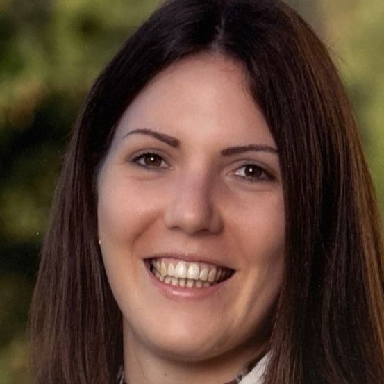 Eine lächelnde Frau mit braunen Haaren und Augen schaut direkt in die Kamera. Sie trägt ein weißes Hemd mit Kragen und scheint im Freien zu stehen, mit einem verschwommenen grünen Hintergrund.