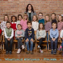 Ein Gruppenfoto einer Klasse von Kindern und ihrer Lehrerin, vor einer Ziegelwand, mit den Worten 'Volksschule Kotschach - Mauthen 2021' am unteren Rand.