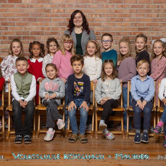 Ein Gruppenfoto einer Klasse von Kindern und ihrer Lehrerin, vor einer Ziegelwand, mit den Worten 'Volksschule Kotschach - Mauthen 2021' am unteren Rand.