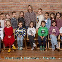 Eine Gruppe von Kindern und einer erwachsenen Frau posiert für ein Foto vor einer Ziegelwand. Sie sitzen alle auf Holzstühlen. Die Kinder tragen verschiedene Kleidungsstücke, und die Frau steht hinter ihnen. Das Bild hat einen Wasserzeichen mit dem Text 'Volksschule Kotschach - Mauthen 2023'.