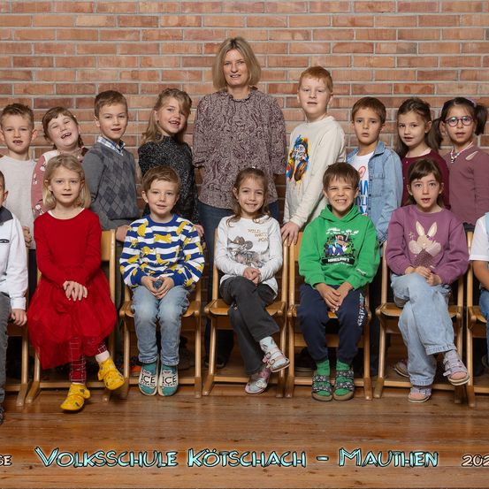 Eine Gruppe von Kindern und einer erwachsenen Frau posiert für ein Foto vor einer Ziegelwand. Sie sitzen alle auf Holzstühlen. Die Kinder tragen verschiedene Kleidungsstücke, und die Frau steht hinter ihnen. Das Bild hat einen Wasserzeichen mit dem Text 'Volksschule Kotschach - Mauthen 2023'.