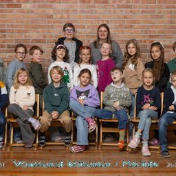Eine Gruppenaufnahme von Kindern und Lehrern an der Volksschule Kötschach-Mauthen. Die Kinder sitzen auf Holzstühlen, mit zwei Lehrern, die hinter ihnen vor einer Ziegelwand stehen.