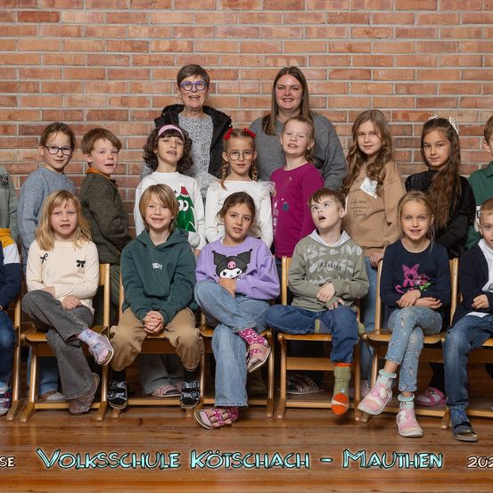 Eine Gruppenaufnahme von Kindern und Lehrern an der Volksschule Kötschach-Mauthen. Die Kinder sitzen auf Holzstühlen, mit zwei Lehrern, die hinter ihnen vor einer Ziegelwand stehen.