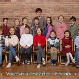 Eine Gruppe von Kindern und einer erwachsenen Frau sitzt vor einer Ziegelwand und lächelt für ein Foto. Sie befinden sich an der Volksschule Kotschach-Mauthen.