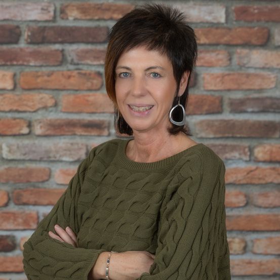 Eine Frau mit kurzem Haar und Ohrringen in einem grünen Pullover posiert mit verschränkten Armen vor einer Ziegelwand.