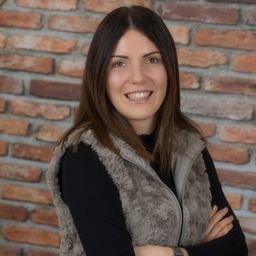 Eine Frau mit braunen Haaren lächelt und steht mit verschränkten Armen vor einer Ziegelwand, sie trägt eine graue Weste über einem schwarzen Langarmshirt.