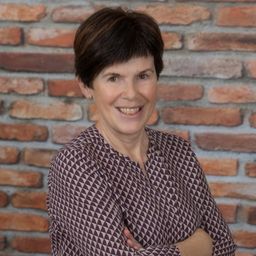 Eine Frau mit kurzen Haaren lächelt und posiert für ein Foto vor einer Ziegelwand in einer gemusterten Bluse.