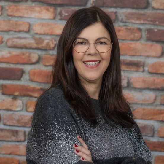 Eine lächelnde Frau mit Brille und grauem Pullover steht mit verschränkten Armen vor einer Ziegelwand.