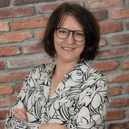 Eine Frau mit kurzen Haaren, die eine Brille und ein gemustertes Hemd trägt, steht lächelnd mit verschränkten Armen vor einer Ziegelwand.