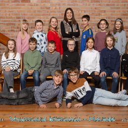 Eine Gruppe von Kindern und einer Frau posiert für ein Foto vor einer Ziegelwand. Die Kinder sitzen und stehen in Reihen, einige tragen eine Brille. Die Frau steht in der Mitte und lächelt. Das Bild hat einen Wasserzeichen mit dem Text 'Volksschule Koetschach - Mauthen 2023'.