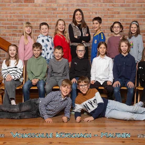 Eine Gruppe von Kindern und einer Frau posiert für ein Foto vor einer Ziegelwand. Die Kinder sitzen und stehen in Reihen, einige tragen eine Brille. Die Frau steht in der Mitte und lächelt. Das Bild hat einen Wasserzeichen mit dem Text 'Volksschule Koetschach - Mauthen 2023'.
