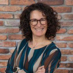 Eine lächelnde Frau mit lockigem Haar, die eine Brille und ein gemustertes Oberteil trägt, steht vor einer Ziegelwand.