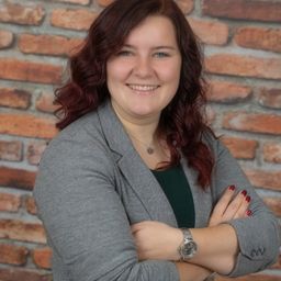 Eine Frau mit roten Haaren und einem grauen Blazer steht selbstbewusst mit verschränkten Armen und lächelt vor einer Backsteinwand.