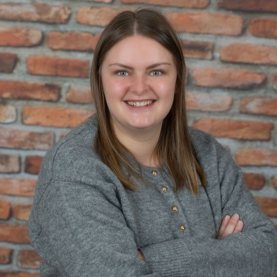 Eine Frau mit langen Haaren und einem grauen Pullover steht mit verschränkten Armen vor einer Ziegelwand und lächelt.