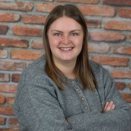 Eine Frau mit langen Haaren und einem grauen Pullover steht mit verschränkten Armen vor einer Ziegelwand und lächelt.