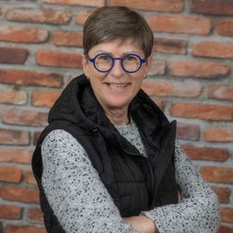 Eine Frau mit einer Brille und einem schwarzen Westchen lächelt mit verschränkten Armen vor einer Ziegelwand.