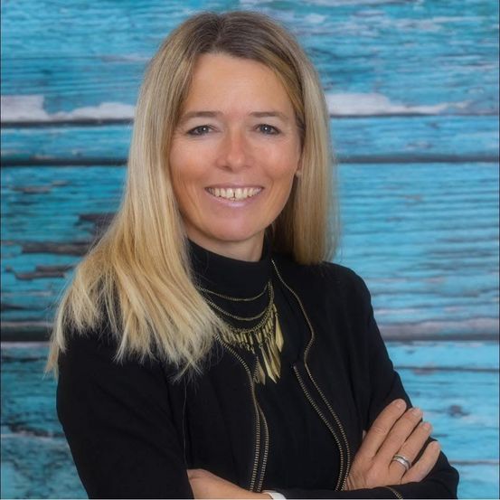 Eine Frau mit blonden Haaren lächelt und posiert für ein Foto vor einem blauen Holzhintergrund. Sie trägt ein schwarzes Rollkragenshirt und eine Halskette.