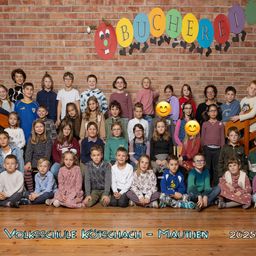 Eine Gruppenaufnahme von Schülern der Grundschule Kotschach-Mauthen im Jahr 2023, in Reihen angeordnet, mit einem bunten Buchstabenbanner darüber.